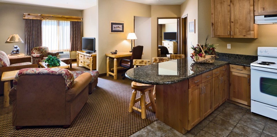 Wide view of a room with a kitchen with an oven and breakfast bar and the living room with sofa, chairs, tv, and desk